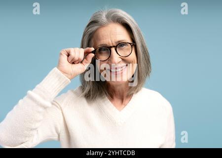 Ritratto di una bella donna matura e positiva che indossa eleganti occhiali da vista, guarda la fotocamera, posa isolata su sfondo blu. Visione, trattamento conc Foto Stock