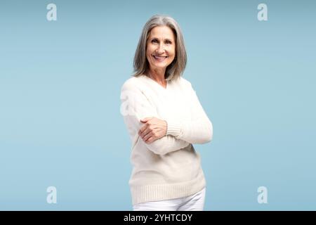 Donna anziana attraente, bella e sorridente che indossa abiti casual, donna d'affari anziana con le braccia incrociate che guarda la fotocamera, posa isolata su blu Foto Stock