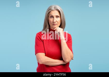 Donna dai capelli grigi, matura e seria, con un maglione rosso che pensa, guarda la fotocamera, posa isolata su sfondo blu. Concetto di pubblicità Foto Stock