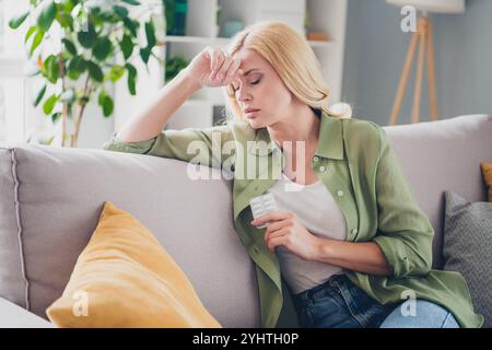 Una donna di mezza età con i capelli biondi si rilassa sul divano a casa, apparendo premurosa e riposante mentre tiene in mano una confezione di farmaci. Foto Stock