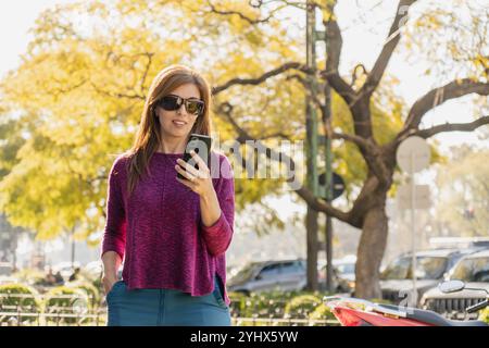 L'elegante donna di mezza età sorride mentre guarda il suo smartphone in un parco al tramonto. Indossando occhiali da sole e un maglione viola, tiene una mano dentro di lui Foto Stock