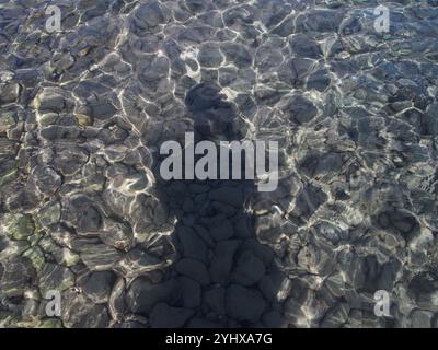L'ombra di una persona gettata sull'acqua poco profonda dell'oceano in una spiaggia di ciottoli a Corfù, in Grecia. Concetto: solo Travel, solitudine, Foto Stock