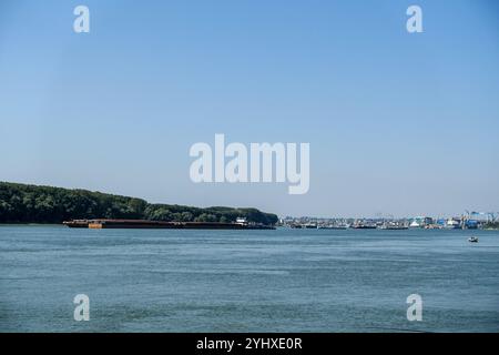 Sponda rocciosa del Danubio/Donau in Romania con lo skyline della città di Galați visibile attraverso le acque calme sotto un cielo limpido e una chiatta di passaggio Foto Stock