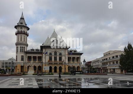 Il Municipio di Buzău, un edificio storico in stile europeo con torre e tetto piastrellato nella piazza della città con bandiere e fontane in una giornata nuvolosa Foto Stock