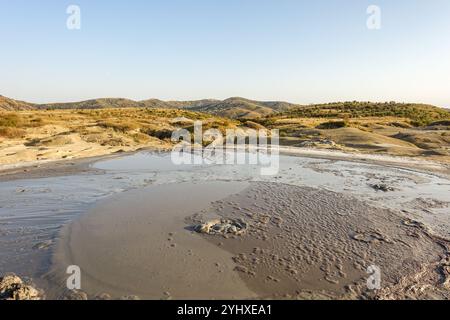 Paesaggio caratterizzato da un vulcano di fango gorgogliante in un terreno arido e asciutto con colline e vegetazione sparsa sotto un cielo azzurro Foto Stock