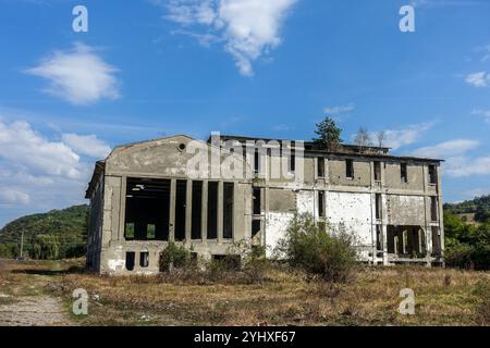 Vecchio edificio industriale abbandonato con finestre rotte e vegetazione sovrastata sotto un cielo blu in un ambiente rurale vicino a Câmpulung, in Romania Foto Stock