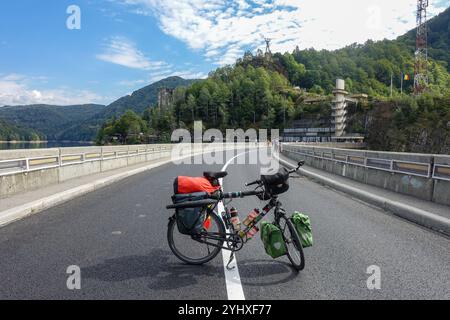 Bicicletta turistica completamente carica parcheggiata sulla diga panoramica di Vidraru, Transfăgărășan, Romania, con colline boscose Foto Stock