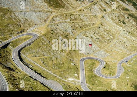 Veduta aerea della tortuosa strada di montagna della Transfagarasan con una funivia rossa che si muove sopra le curve strette e il paesaggio roccioso in una giornata di sole Foto Stock