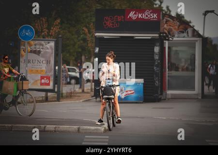 NOVI SAD, SERBIA - 8 SETTEMBRE 2024: Donna in bicicletta mentre utilizza il suo smartphone a Novi Sad, Serbia, che illustra l'intersezione tra technol Foto Stock