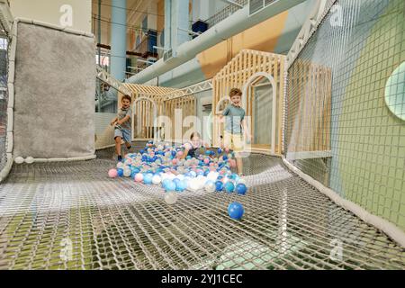 Simpatico ragazzo ridendo che corre alla ricerca di palloncini blu lungo il parco giochi a rete mentre i suoi amici si divertono a giocare sulla rete del parco giochi Foto Stock