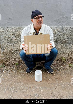 Uomo con un berretto e un bicchierino seduto a terra, tenendo un cartello di cartone con un contenitore bianco per raccogliere soldi davanti, Italia Foto Stock