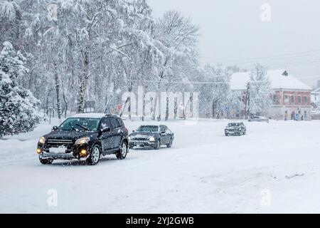 Auto che guidano su una strada scivolosa e innevata su una strada della città durante una forte nevicata la sera in inverno. Interruzioni del traffico dovute a torce di neve e bufera Foto Stock