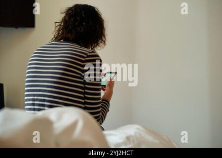 Donna in camicia a righe seduta su un letto con uno smartphone con parete bianca e televisore sullo sfondo. Foto Stock