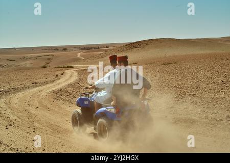 Due persone viaggiano su un quad blu attraverso un paesaggio desertico, lasciando dietro di loro una scia di polvere sotto un cielo limpido, il Marocco Foto Stock