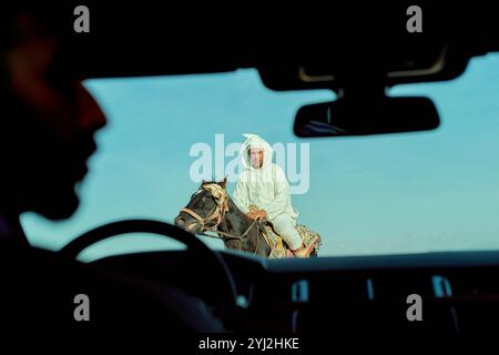 Un uomo in abbigliamento tradizionale cavalca un cavallo, visto attraverso il parabrezza di un'auto con la silhouette di un passeggero in primo piano, il Marocco Foto Stock