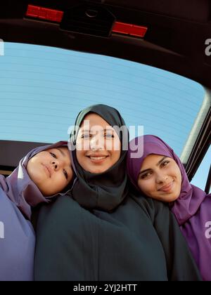 Tre donne sorridenti in hijab posano insieme all'interno di un'auto sotto un cielo azzurro. Foto Stock