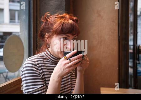 Una donna con i capelli rossi che sorseggia un caffè in un bar, che guarda lontano dalla fotocamera con un'espressione premurosa, Bruxelles, Belgio Foto Stock