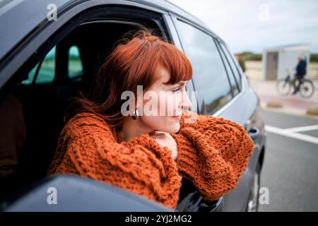 Donna con i capelli rossi che si sporge dal finestrino di un'auto in una giornata nuvolosa e indossa un comodo maglione in maglia arancione. Foto Stock