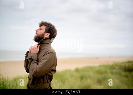 Uomo barbuto in una giacca in piedi su dune erbose che guardano verso il cielo con un'espressione contemplativa, Belgio Foto Stock