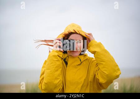 Donna sorridente in impermeabile giallo che tiene in mano una macchina fotografica in una giornata nuvolosa in spiaggia, Belgio Foto Stock