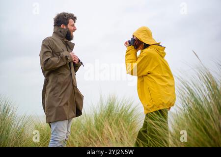 Una persona con un impermeabile giallo scatta una foto di un uomo barbuto sorridente in un cappotto verde contro un cielo nuvoloso, in Belgio Foto Stock