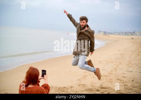 Uomo che salta gioiosamente sulla spiaggia mentre una donna scatta la sua foto con uno smartphone, Belgio Foto Stock