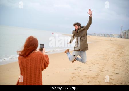 Uomo a mezz'aria che salta su una spiaggia sabbiosa e viene fotografato da una donna con uno smartphone, il Belgio Foto Stock