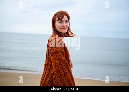 Donna con i capelli rossi in un maglione marrone in piedi su una spiaggia con l'oceano sullo sfondo, Belgio Foto Stock