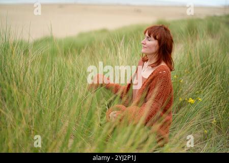 Donna con un maglione arancione bruciato seduta in erba alta con un'espressione serena, Belgio Foto Stock