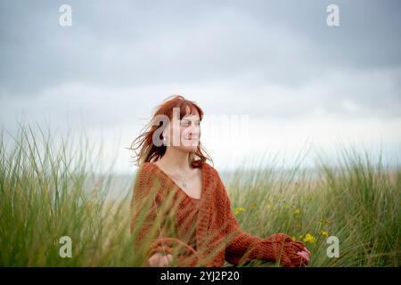 Donna dai capelli rossi in un maglione arancione seduto tranquillamente tra erba alta con un cielo nuvoloso sullo sfondo, Belgio Foto Stock