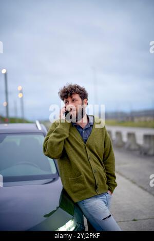 Uomo con capelli ricci che parla al telefono mentre si appoggia a un'auto all'aperto al crepuscolo. Foto Stock