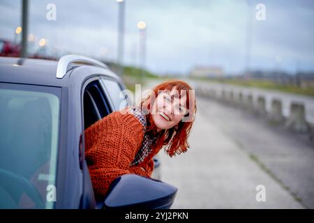 Donna sorridente con capelli rossi che sbirciano fuori dal finestrino di un'auto in una giornata nuvolosa. Foto Stock