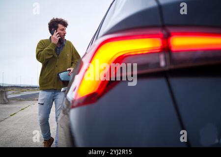 Uomo che parla su uno smartphone accanto a un'auto con luci di coda illuminate in una giornata travolgente, Belgio Foto Stock