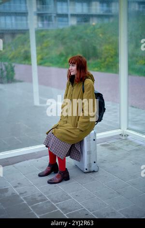 Donna con un cappotto giallo che parla al telefono cellulare mentre è seduta su una valigia presso una fermata dell'autobus con pannelli di vetro, in Belgio Foto Stock