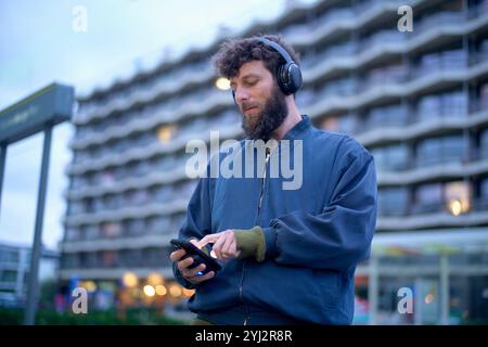 Uomo adulto con capelli ricci che indossa le cuffie con uno smartphone con edifici cittadini sullo sfondo al crepuscolo, Belgio Foto Stock
