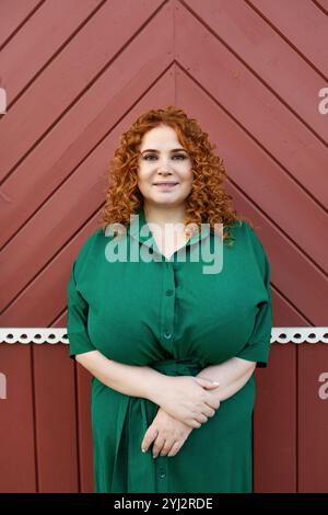 Portrait Plus size bella donna con capelli ricci rossi in abito verde che guarda la fotocamera su sfondo rosso colorato della parete, bellezza e sicurezza Ou Foto Stock