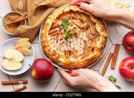 Una persona sta mettendo con cura una galette di mele appena sfornata su un tavolo circondato da mele affettate, bastoncini di cannella e fiocchi di mandorle. Il tiepido kitch Foto Stock