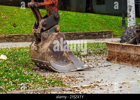 Grande secchio da escavatore sul marciapiede con foglie autunnali sparse in un parco verde. Strumento industriale in un ambiente naturale, che rappresenta il paesaggio urbano Foto Stock