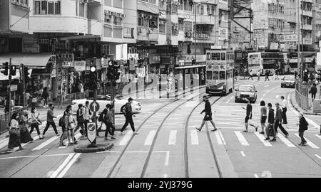 I pedoni percorrono una strada vivace piena di tram, negozi e semafori in una tipica giornata a Hong Kong. Foto Stock