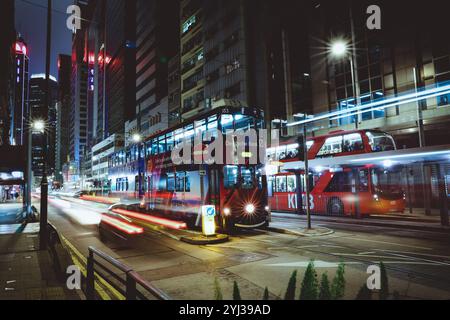 Un vivace tram viaggia lungo una strada trafficata di Hong Kong, illuminata dalle luci della città e dal bagliore dei grattacieli di notte. Foto Stock