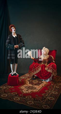 Giovane ragazzo, servitore di corte in parrucca rossa accanto a una donna anziana, imperatrice medievale e cantando per lei. Foto Stock