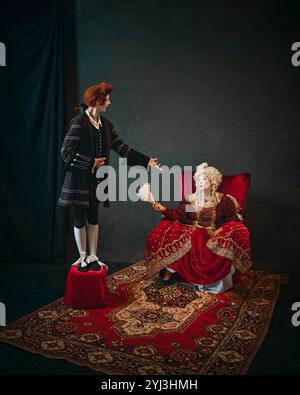 Giovane uomo, servitore di corte in parrucca rossa accanto a una donna anziana, imperatrice medievale e cantando per lei. Foto Stock