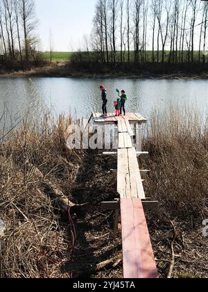 Una famiglia trascorre del tempo di qualità su un molo in legno vicino al lago, circondato dalla natura. Trascorrono una tranquilla giornata all'aperto, con un vivace paesaggio primaverile Foto Stock