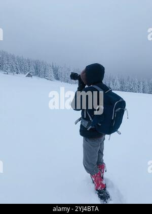 Un fotografo dedicato con uno zaino cattura la bellezza serena di un paesaggio invernale innevato. La scena è tranquilla, con alti alberi innevati Foto Stock