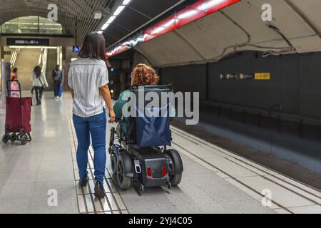 Vista posteriore di una donna caucasica disabile in sedia a rotelle e di un amico asiatico che cammina lungo la stazione della metropolitana Foto Stock