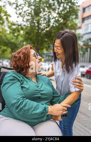 Foto verticale di una donna caucasica felice in sedia a rotelle che riceve il sostegno di un'amica multietnica che cammina insieme per la città Foto Stock