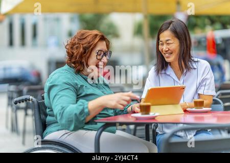 Donna con disabilità che usa un tablet accanto a un'amica asiatica seduta in una caffetteria all'aperto Foto Stock