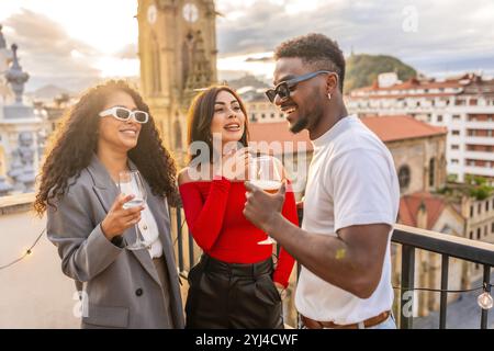 Amici multietnici che chiacchierano e bevono vino sul tetto durante il tramonto con vista sulla città Foto Stock