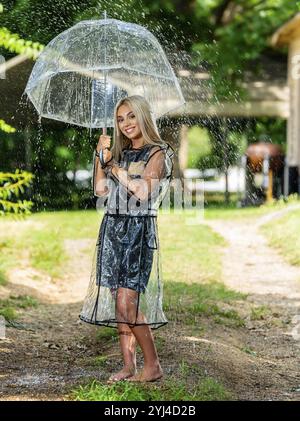 Una radiosa modella bionda posa graziosamente sotto la pioggia, indossando un impermeabile e tenendo in mano un ombrello, il suo sorriso aggiunge fascino alla scena primaverile Foto Stock