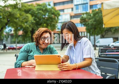 Donna disabile in sedia a rotelle elettrica e amica cinese che usa un tablet seduto in una caffetteria sul marciapiede Foto Stock